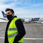 Man at airport runway wearing a reflective vest and face mask near LOT Polish Airlines plane.